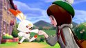 Een meisje met een grijze jas en een groen petje geeft een vuistje aan een wit Pokémon met oranje accenten.