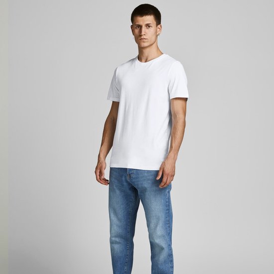 Man in een wit T-shirt en blauwe spijkerbroek, staand, licht gedraaid.