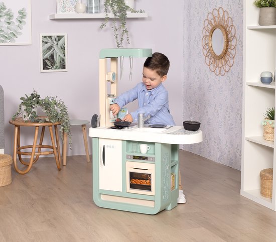 Een jongen in een blauw shirt speelt met een mintgroene speelkeuken met een witte wasbak, kookplaat en oven.