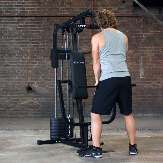 Man met krullend haar, grijs T-shirt en zwarte korte broek doet een oefening aan het Focus Fitness Unit 2 krachtstation.