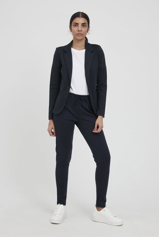 Een vrouw met donker haar draagt een marineblauwe blazer en bijpassende broek met touwtjes. Ze draagt een witte t-shirt en witte sneakers.