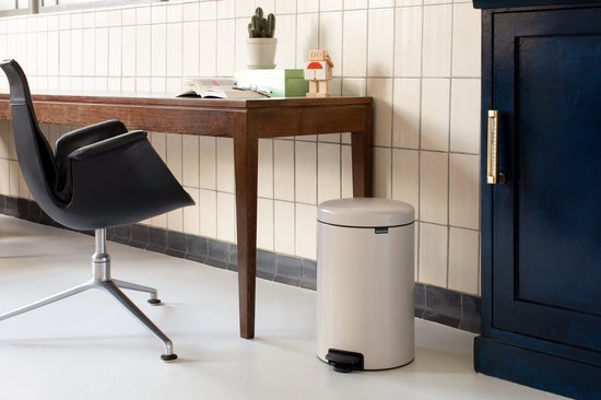 Crèmekleurige Brabantia NewIcon prullenbak van 20 liter naast een houten bureau met een zwarte bureaustoel.