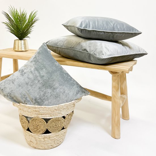 Lichtgrijs velours sierkussen in een rieten mand, naast een houten bankje.