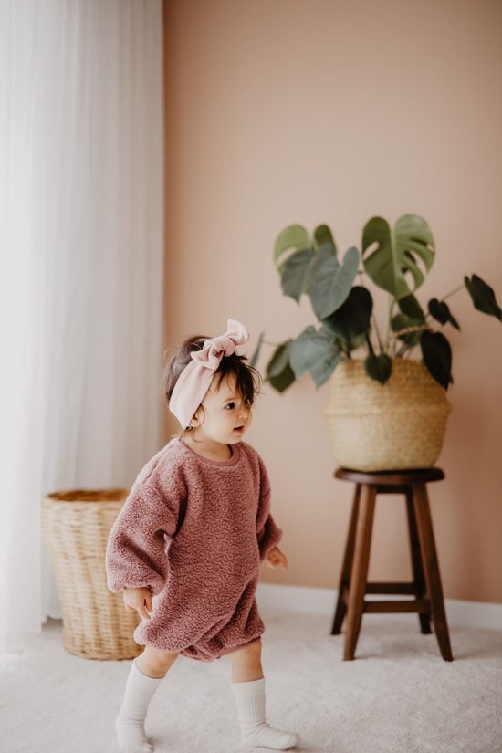 Peuter draagt een roze teddy romper met lange mouwen, gecombineerd met een bijpassende roze hoofdband en witte sokken.