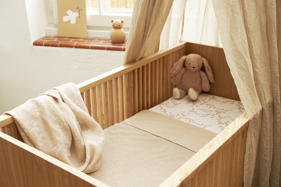 Houten wieg met een bruine konijnenknuffel en een deken met bladerpatroon.