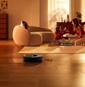Zwarte robotstofzuiger zuigt sporen op een houten vloer, terwijl een moeder en dochter op de achtergrond spelen.