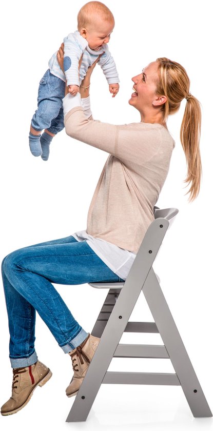 Vrolijke blonde vrouw in beige trui en spijkerbroek tilt lachende baby omhoog