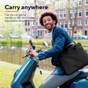 Een man met een zwarte laptoptas om zijn schouder, zittend op een scooter, met een waterweg en gebouwen op de achtergrond.