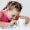 Meisje met vlechten en oorbellen schildert een mok met Stitch; potjes met verf staan ernaast.
