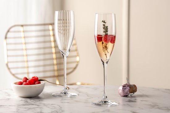 Twee champagneglazen op een marmeren tafel; één leeg, één gevuld met roze drank, frambozen en takjes.