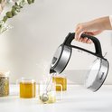 Hand giet kokend water uit glazen waterkoker in theekopjes, rook stijgt op.