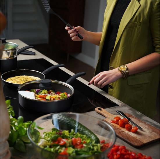 Une personne portant un blazer vert olive cuisine des légumes dans une poêle noire avec un couvercle en verre.