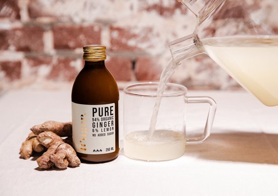 Een bruine fles G'nger biologisch gembersap, 94% gember, 6% citroen, zonder toegevoegde suiker, wordt in een glas gegoten. Naast de fles liggen gemberwortels.