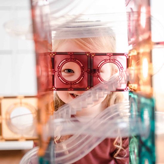 Een jong meisje met blond haar kijkt door een rood Connetix-speelraam, glimlachend naar de camera.