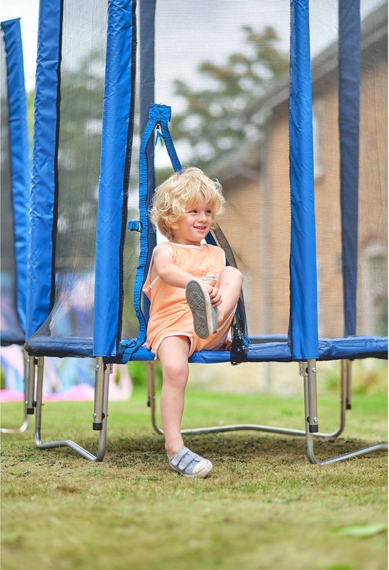 Jongetje met blond haar zit op de rand van een blauwe Plum trampoline en trekt zijn schoen aan.