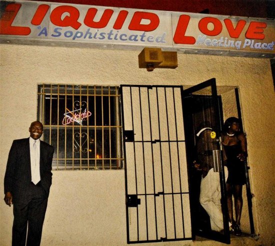 Een man in een zwart pak en witte das staat voor het etablissement 'Liquid Love A Sophisticated Meeting Place'. Twee personen staan in de deuropening.