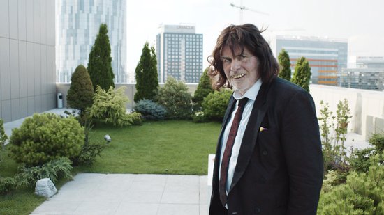Een man met lang bruin haar, een zwart pak en een stropdas staat op een dakterras met groen en gebouwen op de achtergrond.