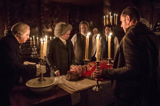 Een groep mannen rondom een liggend lichaam in een kaarsverlichte kamer.