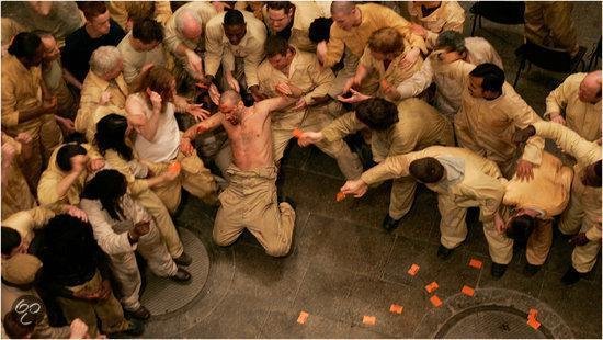 Een groep gevangenen in gele uniformen omringt een man die op zijn knieën zit, met oranje briefjes op de grond.