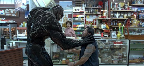 Venom, een groot zwart wezen met witte ogen en tanden, grijpt een man met tatoeages in een winkel.