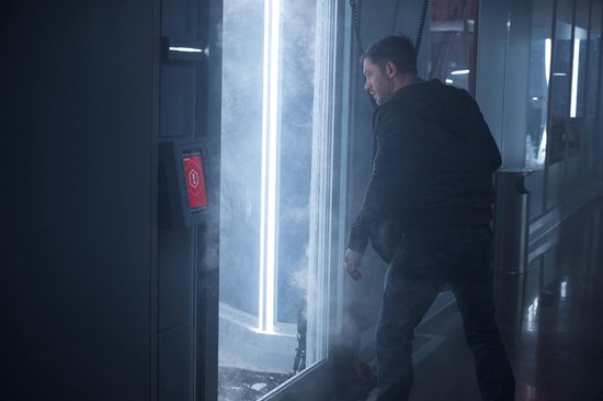 Een man in een zwart jack en spijkerbroek kijkt naar een futuristische deur met blauw licht en een rood waarschuwingssymbool.