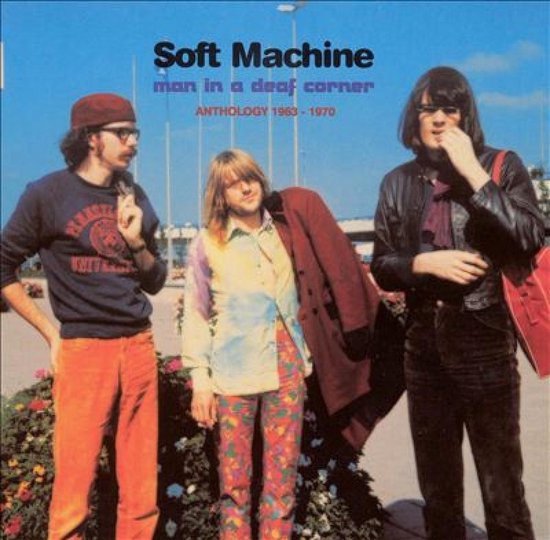 Soft Machine, Man in a Deaf Corner, Anthology 1963 - 1970. Drie mannen staan voor bloemen. Een man links draagt een bordeauxrode baret, bril en donkerblauwe trui. Een man in het midden draagt een kleurrijke blouse en broek en een jas over zijn schouder. Een man rechts draagt een leren jas, sjaal en een rode tas.
