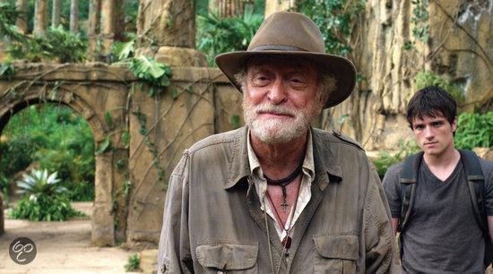 Een oudere man met een hoed en baard, in een safari-outfit, met een jongere man op de achtergrond.