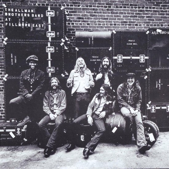 De Allman Brothers Band, zwart-wit, staand en zittend, voor een bakstenen muur met grote koffers met de tekst 'The Allman Brothers Band at Fillmore East'.