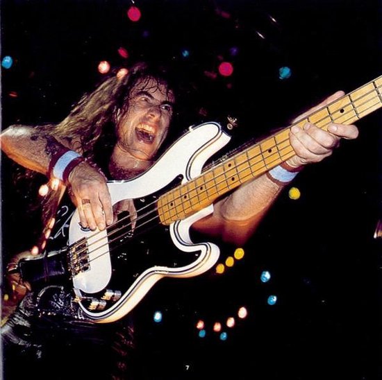 Een bassist met lang bruin haar speelt op een witte basgitaar met zwarte slagplaat, verlicht door gekleurd licht.