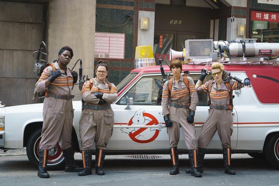 Vier vrouwen in Ghostbusters-uniformen die buiten voor een oude ambulance staan.