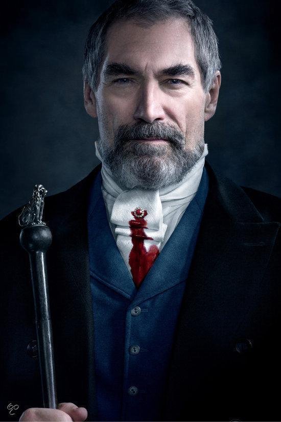 Een man met grijze baard, draagt een blauwe vest en zwarte jas. Witte das met bloedvlek.