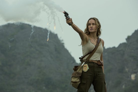 Brie Larson als Mason Weaver, met een pistool dat rook en vuur afschiet, in een groen, rotsachtig landschap.