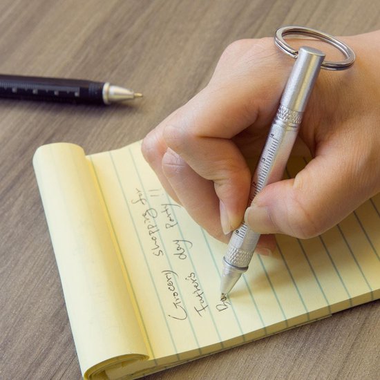 Hand met zilveren multitool pen die in een notitieboekje schrijft. Naast de pen ligt een zwarte multitool pen.