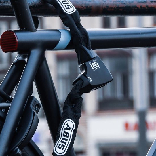 Zwart ABUS kettingslot, bevestigd aan een fietsframe in een stedelijke omgeving. Twee ABUS sleutels zijn zichtbaar.