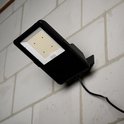 Zwarte LED-schijnwerper met LED's aan, gemonteerd op een bakstenen muur.
