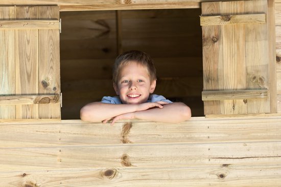 Jongetje met blond haar, lachend, leunt op een houten speelhuisje.