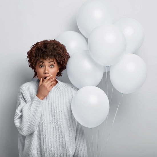 Verraste vrouw met afro-kapsel in lichtblauwe trui, hand voor de mond, met een bos witte ballonnen.