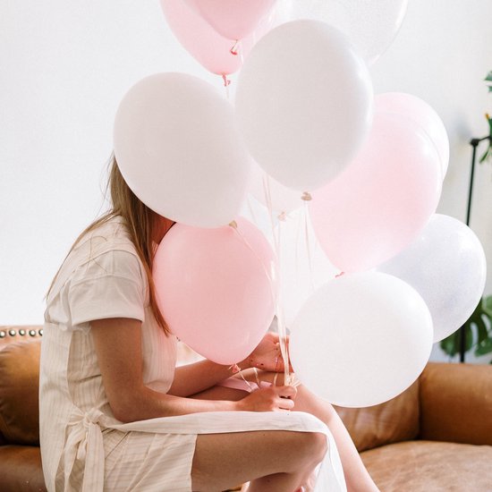 Vrouw in beige jurk zit op een bruine bank, omringd door pastelroze en witte ballonnen.