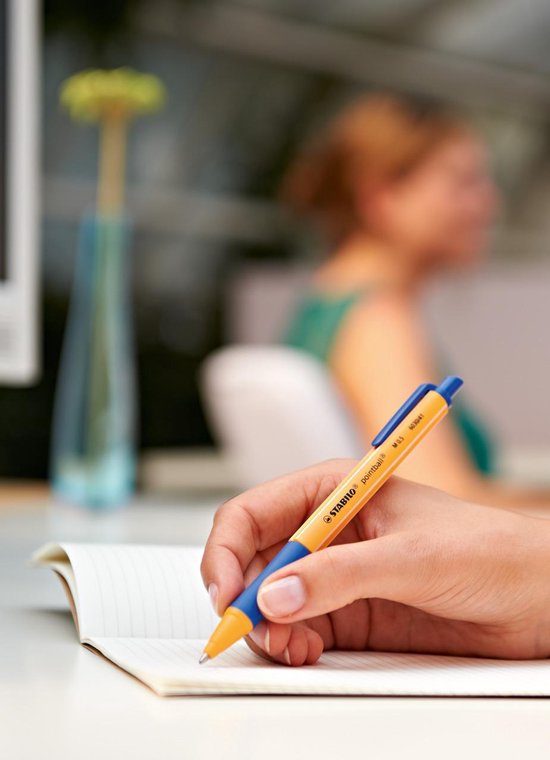 Hand die met een oranje STABILO pointball balpen met blauwe grip in een notitieboekje schrijft.