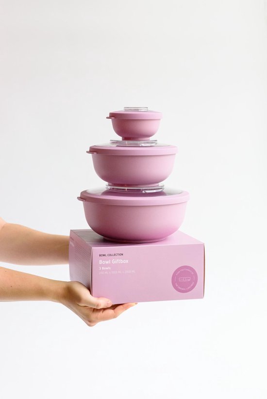 Une boîte cadeau rose avec des bols roses empilés dessus, tenue par deux paires de mains.