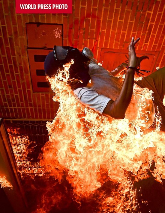 Een persoon in vlammen gehuld, met een gasmasker en beschermende kleding, staand tegen een bakstenen muur.