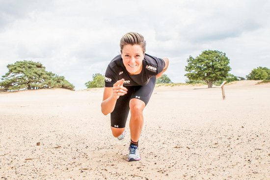 Vrouw in sportkleding, een zwarte compressiebroek en shirt, rent in het zand met bomen op de achtergrond.