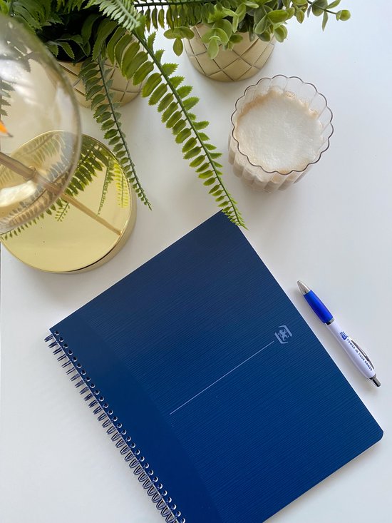 Donkerblauw A4+ notitieboek met spiraalbinding, naast een pen en planten op een bureau.