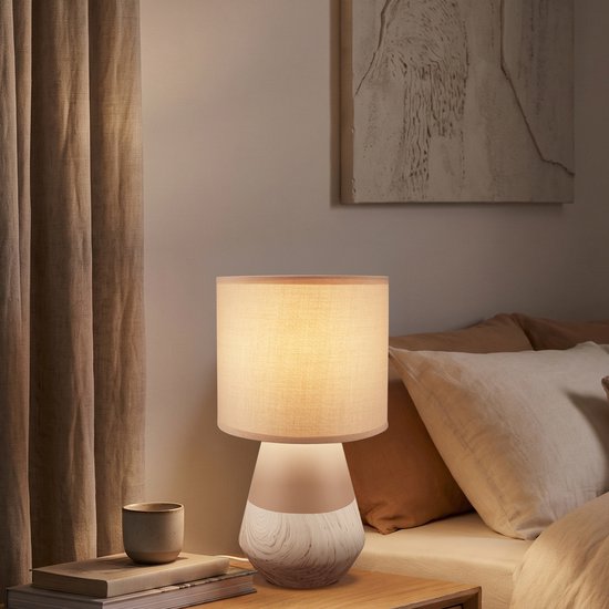 Tafellamp met beige stoffen kap en een voet van keramiek met houtnerf-look, naast een bed.