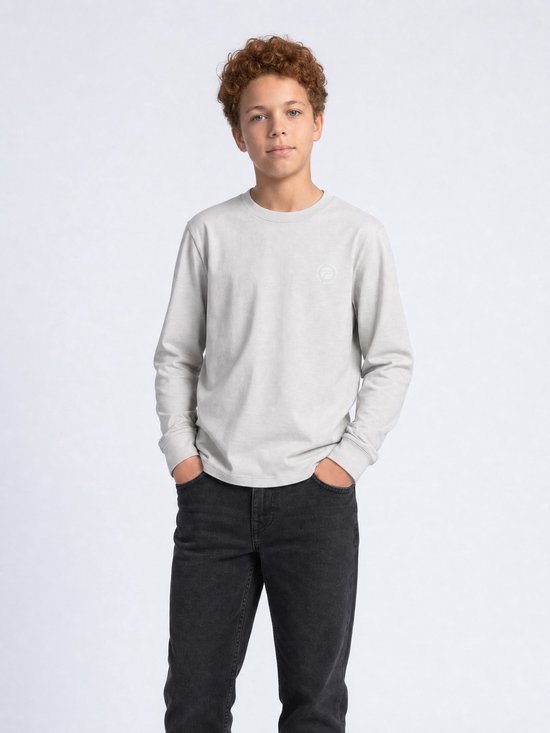 Jongen met krullend rood haar, handen in de zakken van een zwarte jeans en een lichtgrijze longsleeve met logo.