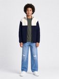 Model draagt een donkerblauw en off-white teddyjack met rits, gecombineerd met een olijfgroen shirt en gescheurde jeans.