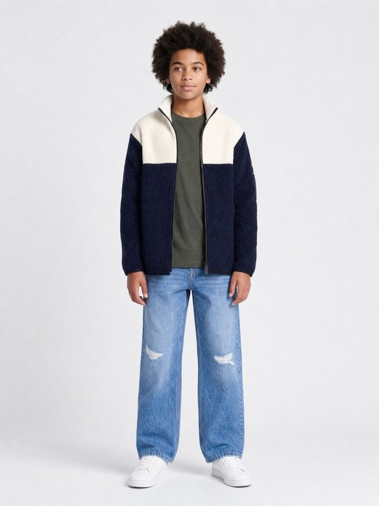 Model draagt een donkerblauw en off-white teddyjack met rits, gecombineerd met een olijfgroen shirt en gescheurde jeans.