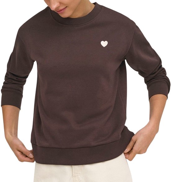Model draagt bruine sweater met witte hart-applicatie, met handen aan de zoom.
