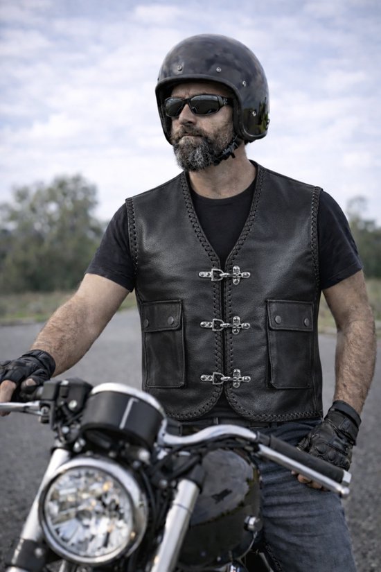 Man met baard draagt een zwart leren bikerjack, helm en zonnebril, leunend op een motorfiets.