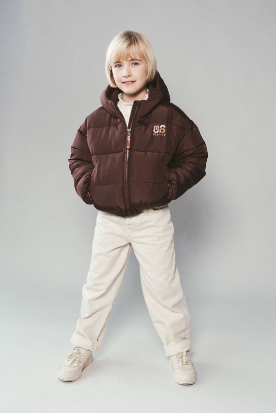 Kind in een bruine pufferjas met capuchon, crème kleurige broek en lichte sneakers.